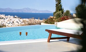 A pool with two glasses of a beverage, a wooden bench, and a view of a coastal town and mountains. - Dipolis Apartments (Syros)