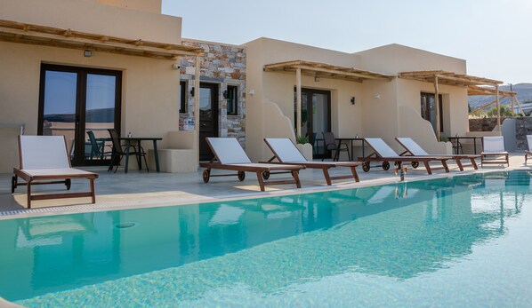 A poolside area with lounge chairs and tables, a clear pool, and a stone facade building. - Dipolis Apartments (Syros)