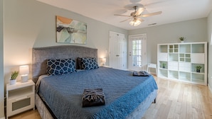 A bedroom with a large bed, a ceiling fan, a nightstand with a lamp, a white wardrobe, and a painting on the wall.