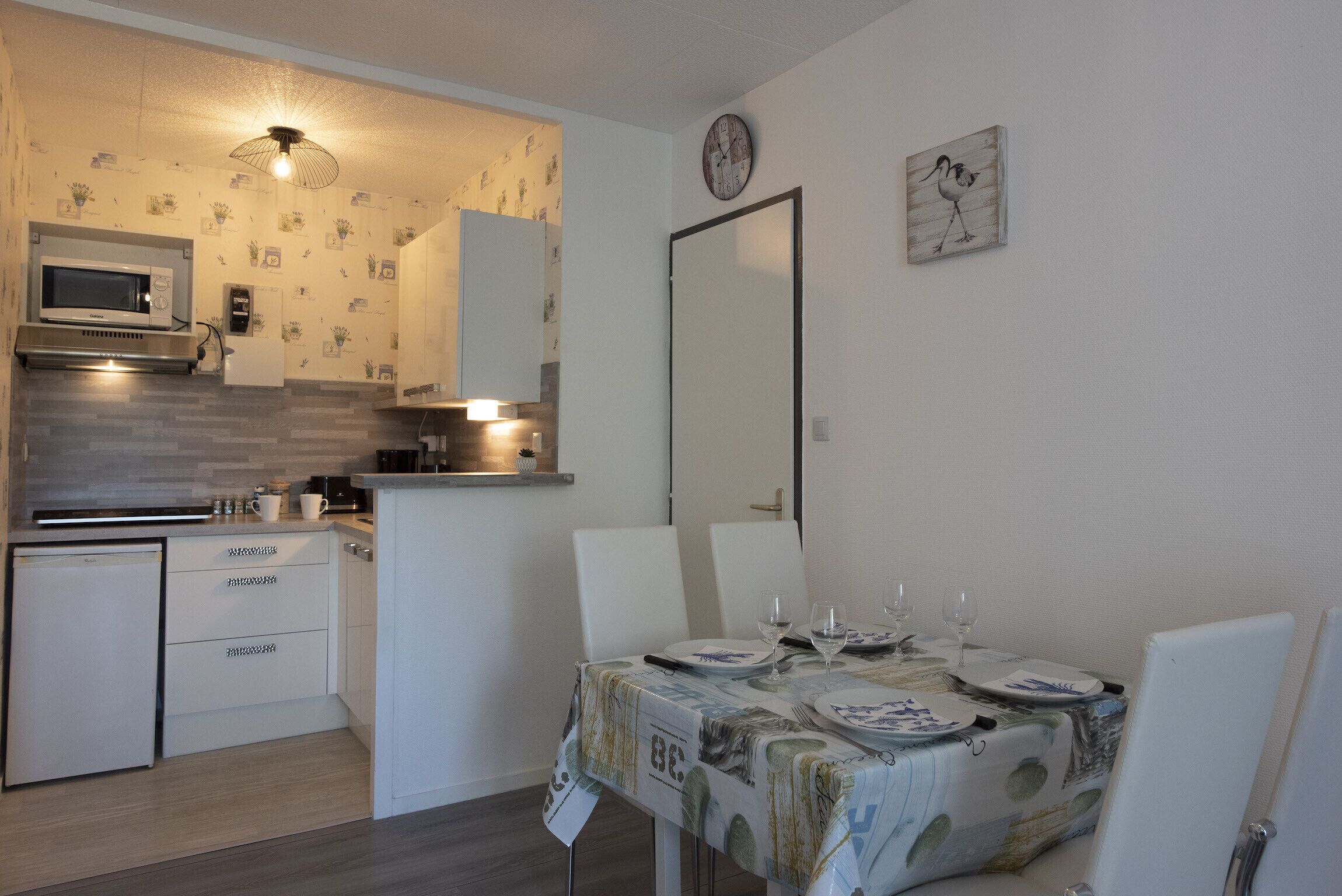 A compact kitchen with a microwave, sink, and cabinets. A dining table with chairs is set for four, and a clock is on the wall.
