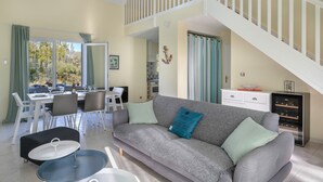 A modern living room with a grey sofa, dining table, and chairs. A staircase leads to an upper level.
