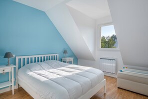 Et soveværelse med skråvæg, stort vindue, hvid seng, natbord med lampe og radiator.