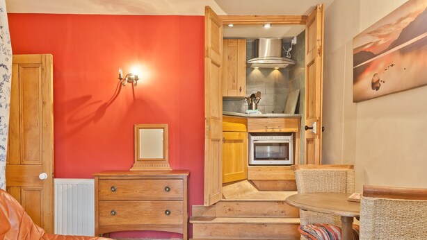 A compact living space with a kitchenette, a dining area with wicker furniture, and a red accent wall. - Lake District village apartment with private riverside hot tub (Coniston)