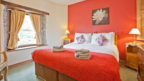 A bedroom with a red wall, a bed with white linens, two pillows, a red blanket, and a wooden nightstand with a lamp. - Lake District village apartment with private riverside hot tub (Coniston)