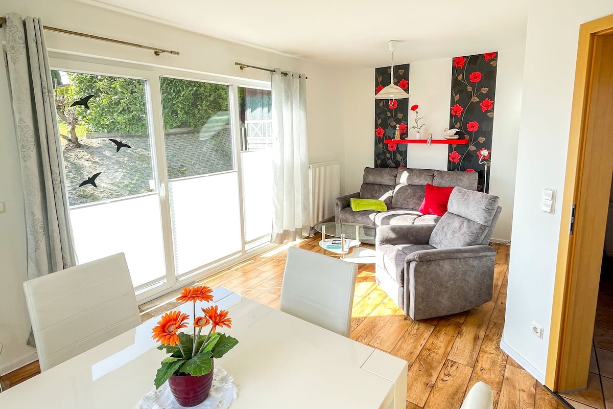 A modern living room with a grey sofa, a dining table with a vase of flowers, and a view of the outdoors through large windows.