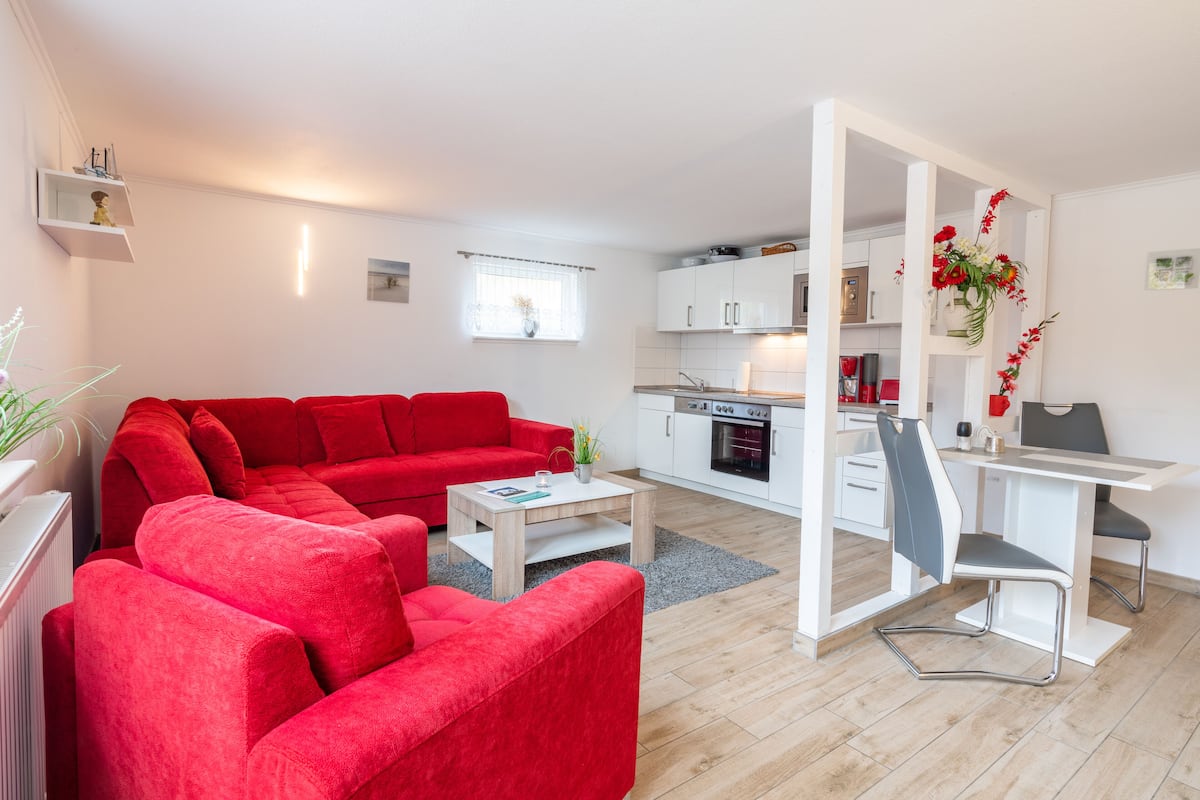 A modern living room with a red sofa, a small coffee table, and a kitchen area with white cabinets and a dining table.