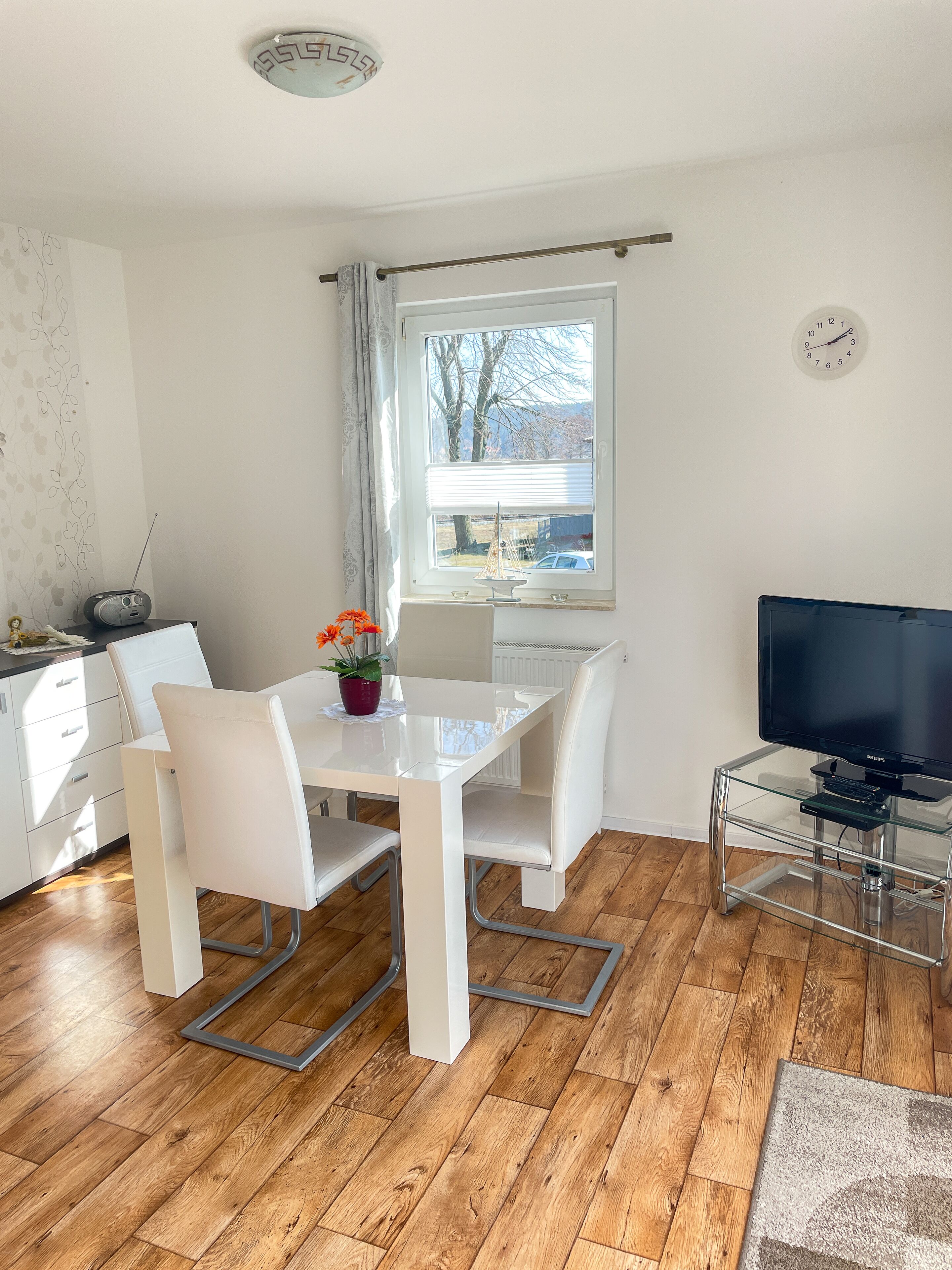 Una sala de estar moderna con una mesa de comedor, sillas blancas, una mesa de centro de vidrio, un televisor de pantalla plana y piso de madera.