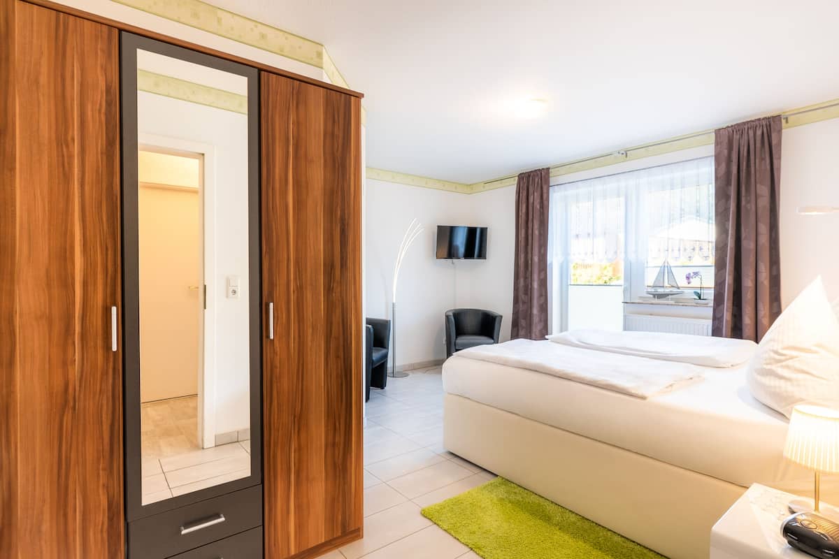 A modern hotel room with a large wardrobe, a bed, a desk with a chair, a TV, and a window with curtains.