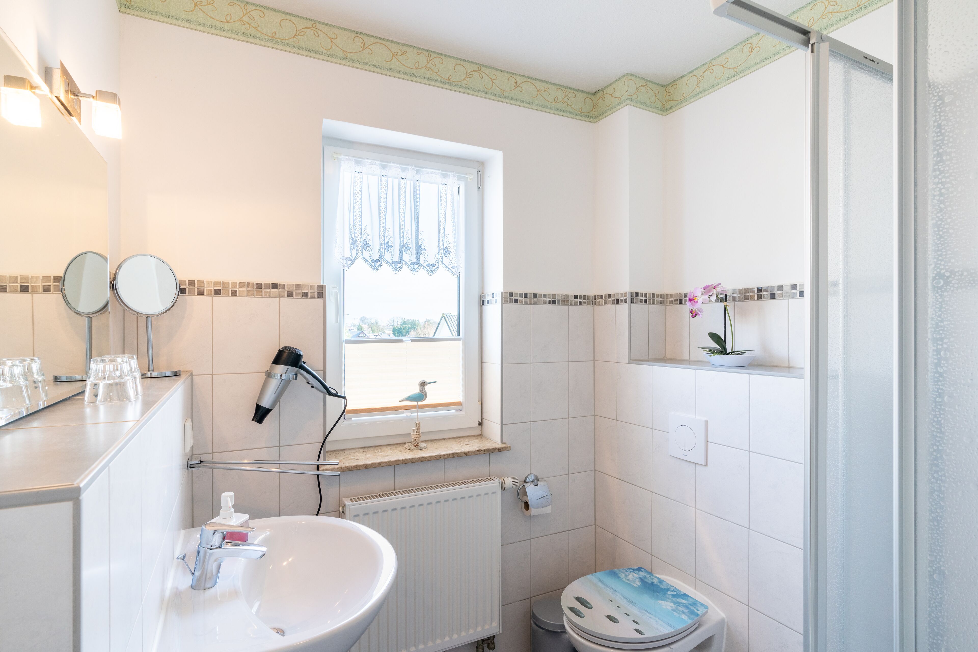 Une salle de bain comprenant un lavabo, deux miroirs, une fenêtre avec un rideau, un radiateur, des toilettes et un espace douche.