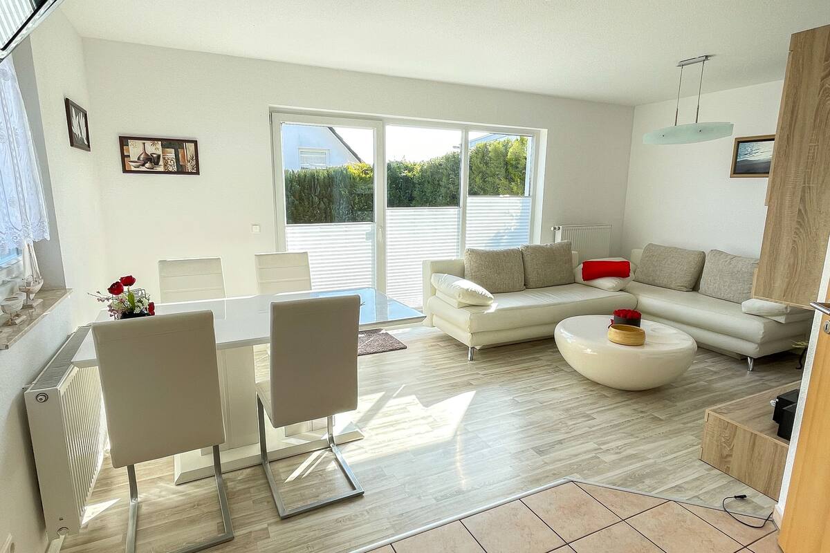 A modern living room with a white sofa, a dining table with chairs, and a large window with blinds.