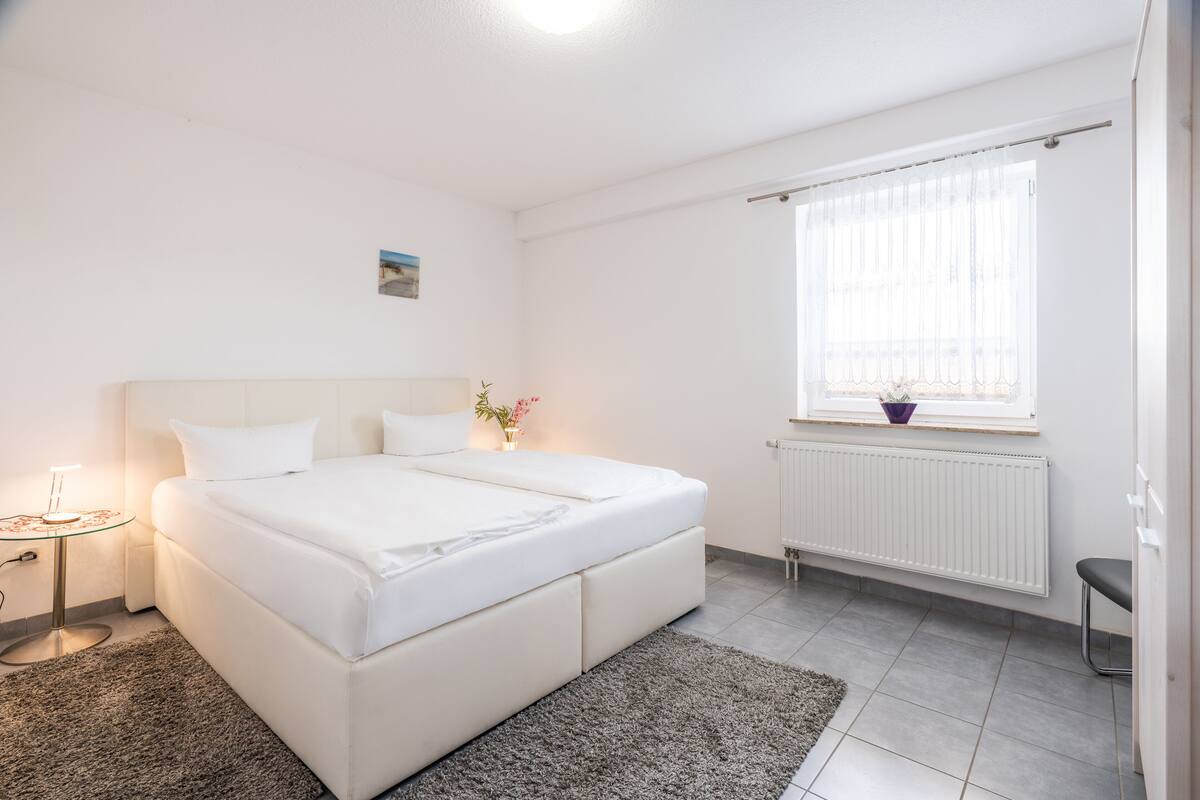 A modern bedroom with a large bed, a bedside table with a lamp, a window with sheer curtains, and a radiator.