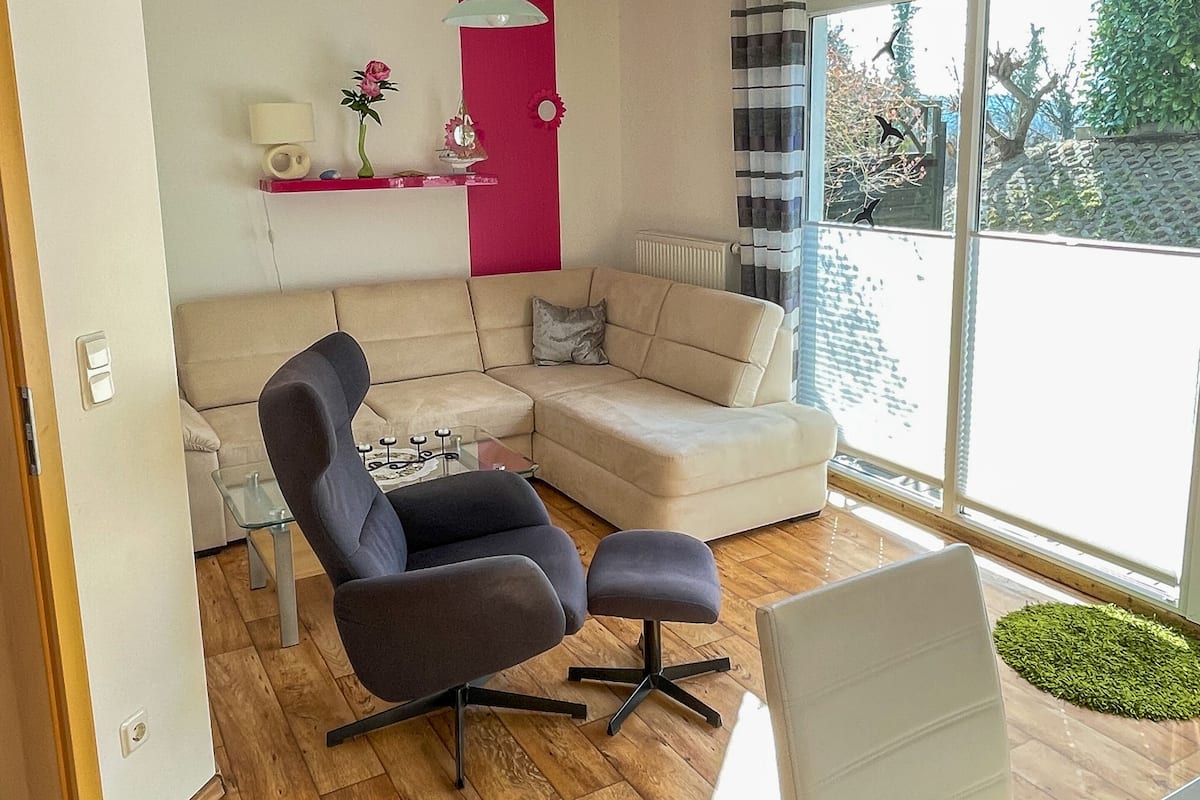 A modern living room with a beige sofa, a dark armchair, and a glass coffee table. A red accent wall and a window with a view of trees are visible.