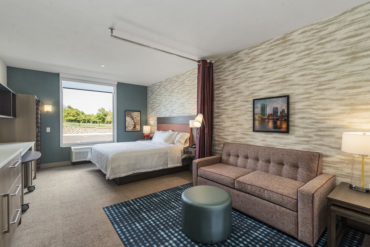 Une chambre d’hôtel avec un lit, un canapé, un bureau et une grande fenêtre donnant sur un paysage verdoyant.