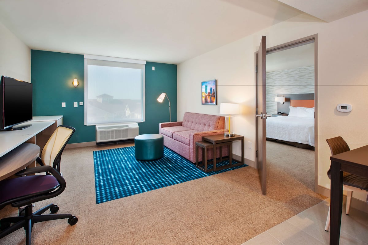Une chambre d’hôtel équipée d’un bureau, d’une chaise, d’un canapé, d’une table de chevet, d’une lampe et d’un lit agrémenté d’un tableau représentant un paysage urbain.