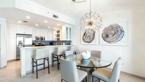 A modern kitchen with white cabinetry, a central island, and a dining area with a glass table and chairs. - Beachcomber 21 by Grand Cayman Villas (7 Mile Beach)