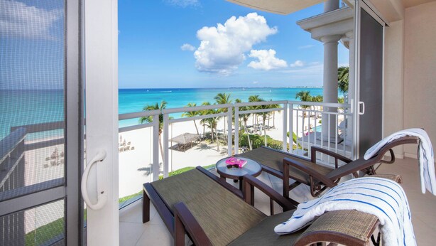 A balcony with a view of the beach, lounge chairs, and a table with a pink item on it. - Beachcomber 21 by Grand Cayman Villas (7 Mile Beach)