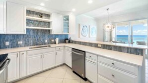 A modern kitchen with white cabinets, a stainless steel dishwasher, and a blue backsplash. - Beachcomber 21 by Grand Cayman Villas (7 Mile Beach)