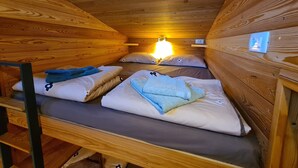 Tunnelmallinen mökkihuone, jossa on puuseinät ja kalteva katto. Huoneessa on kerrossängyt, joissa on tyynyt ja peitot, pieni hylly ja seinään kiinnitetty valaisin.