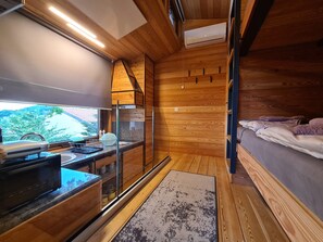 L'intérieur d'un chalet moderne comprend un coin cuisine, une grande fenêtre donnant sur un paysage verdoyant et un espace nuit confortable avec un lit et des oreillers.