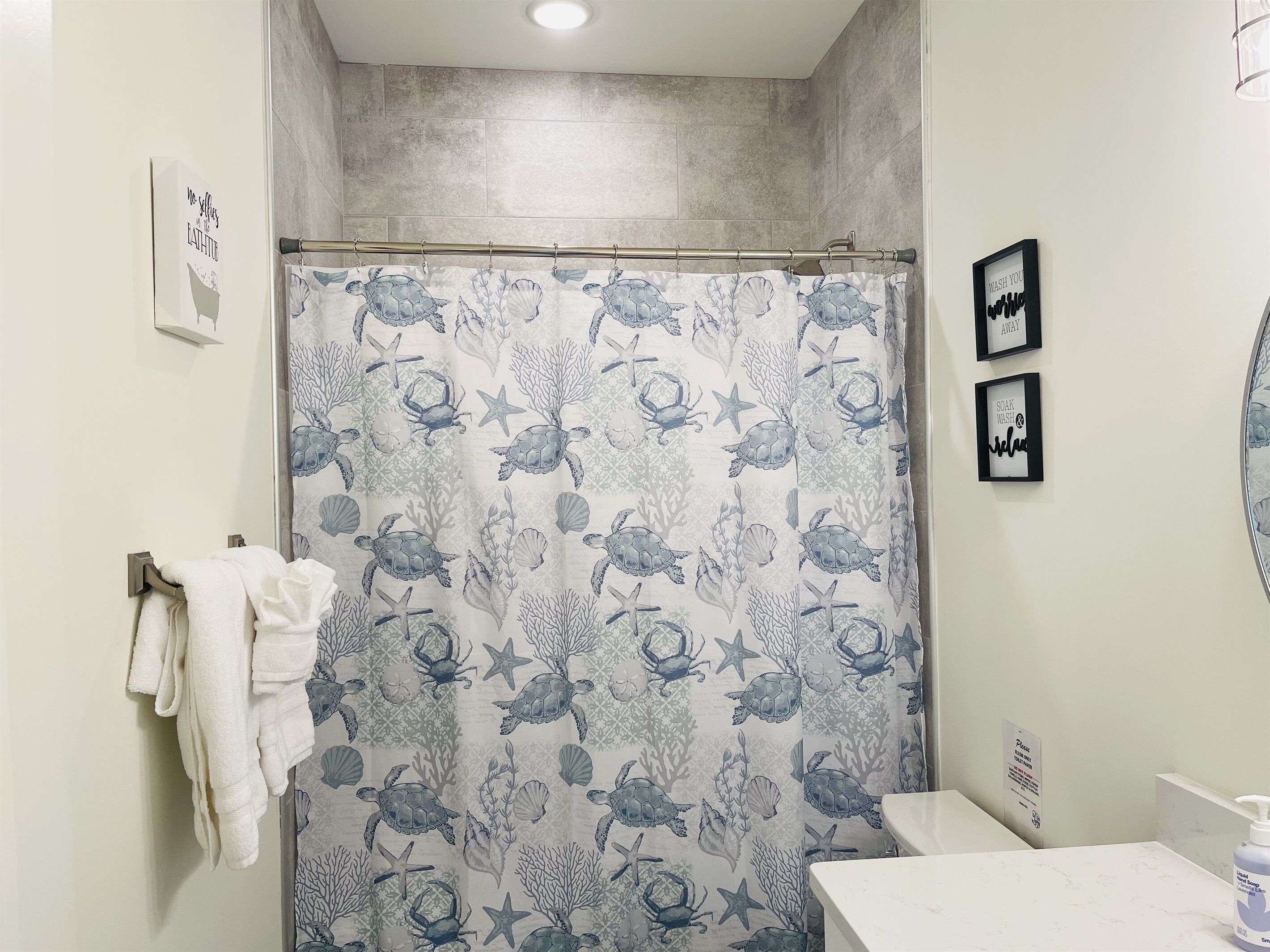 A bathroom with a shower curtain featuring sea turtle and coral patterns, a towel rack with a white towel, and two framed signs on the wall.