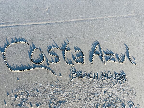 The words "Costa Azul" and "Beach House" written in the sand.