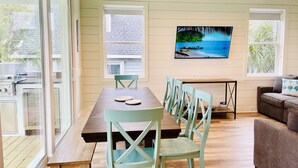 Un coin repas avec une table en bois et des chaises bleu clair, un tableau représentant une scène de plage accroché au mur, et une vue à travers la porte vitrée.