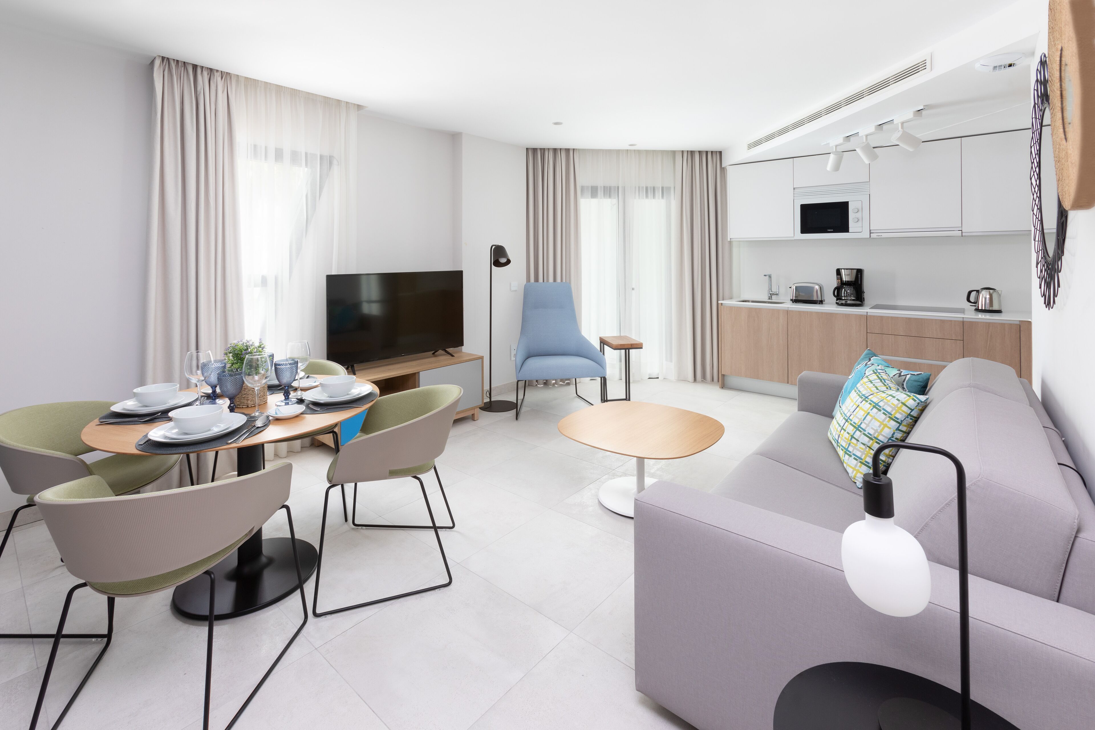 A modern living room with a sofa, dining table, and chairs. There is a television, a coffee machine, and a microwave in the kitchen area.