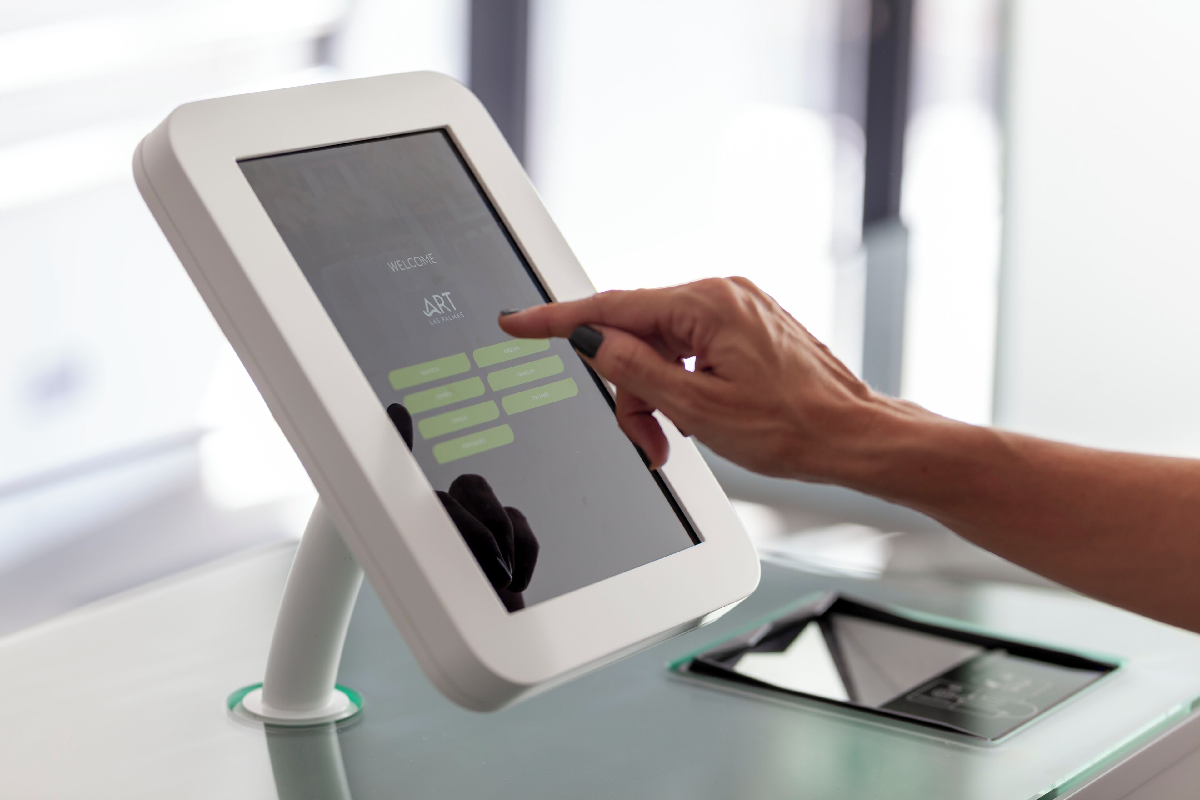 Un quiosco digital con pantalla táctil, una mano interactuando con él y una tableta sobre un soporte cercano.