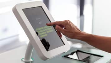 Un kiosque numérique doté d’une interface à écran tactile, une main interagissant avec celui-ci, et une tablette posée sur un support à côté.