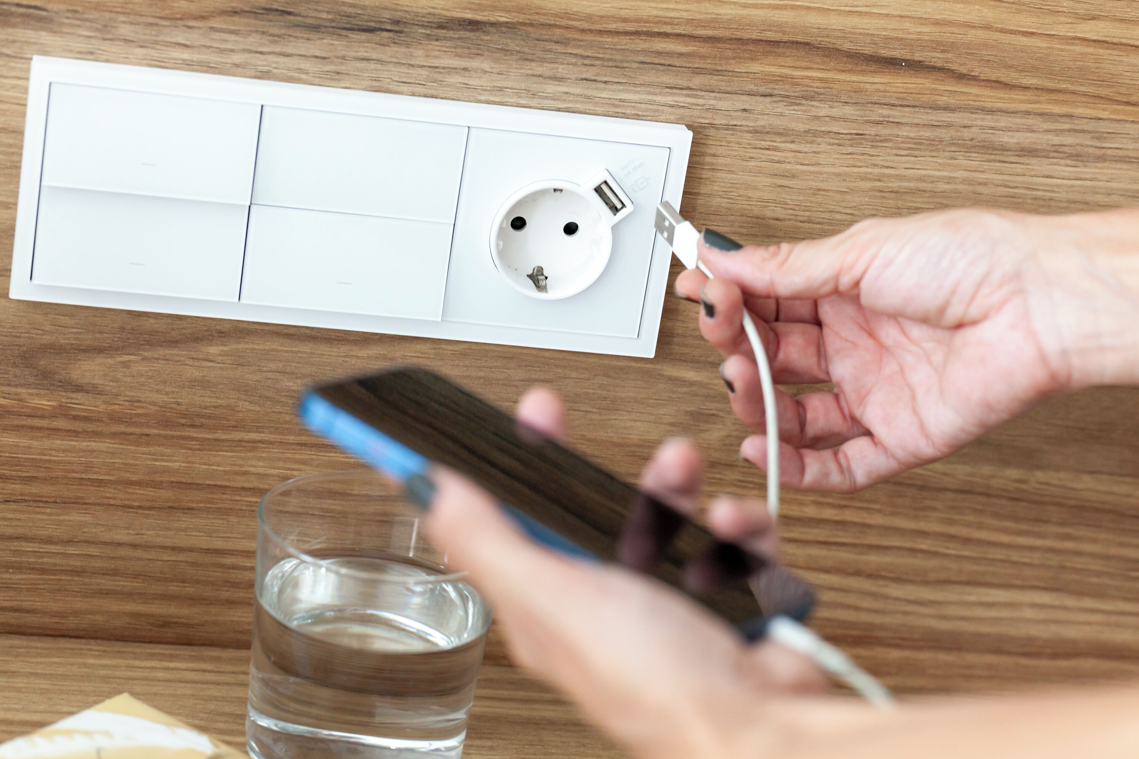 A hand plugging a USB charger into a white electrical outlet on a wooden surface.