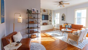 Una sala de estar con un sofá de cuero marrón, un sillón blanco, una mesa de centro y una estantería.