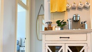 Un mueble blanco de madera con puertas de cristal, una cafetera, un azucarero y una toalla amarilla colgada.