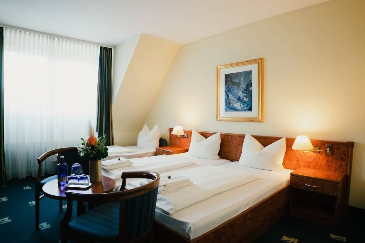 A hotel room with two beds, a desk, a chair, a table with a vase of flowers, and a painting on the wall.