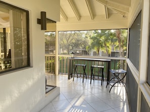 A balcony with a table and chairs, a view of trees and a fence, and a window with curtains.