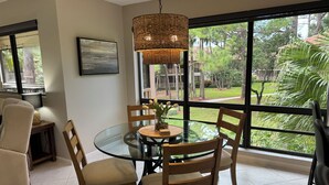 Zona pranzo con tavolo in vetro, sedie in legno, lampada a sospensione in rattan e vista su un rigoglioso giardino attraverso ampie finestre.