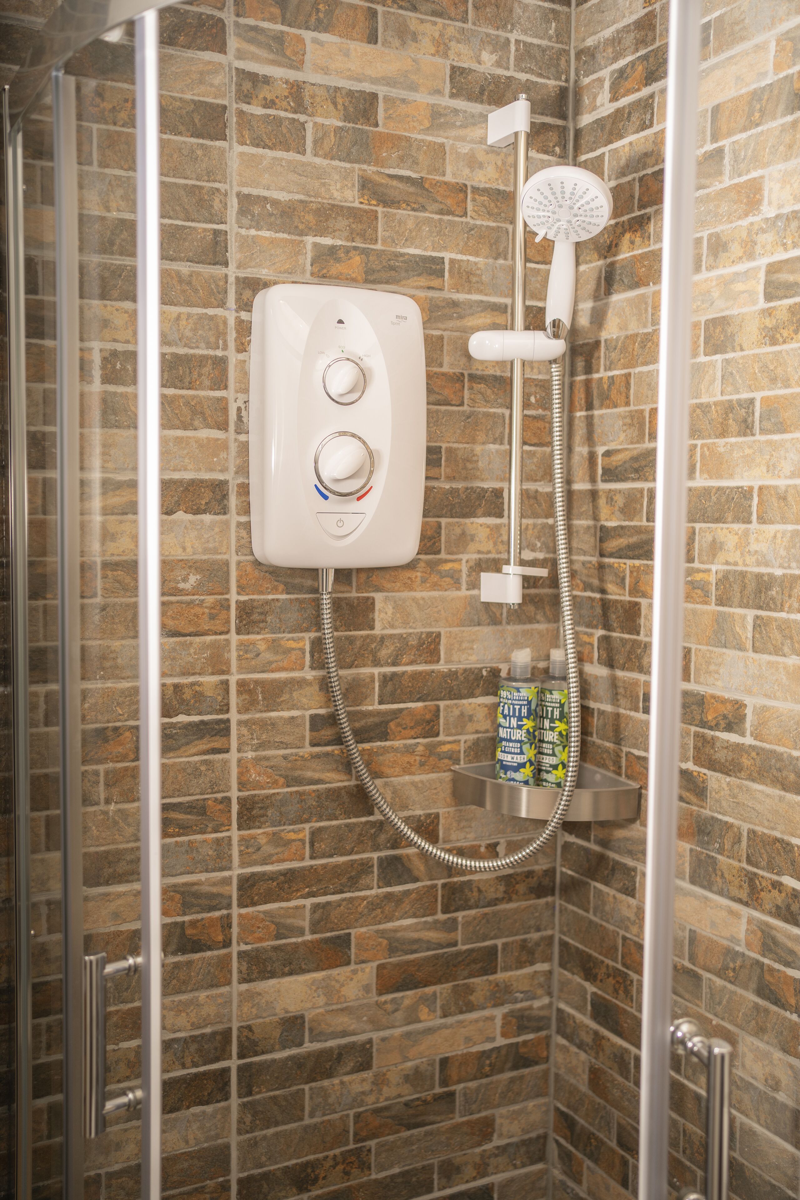 A shower with a control panel, a handheld showerhead, and a bottle of shampoo on a shelf.