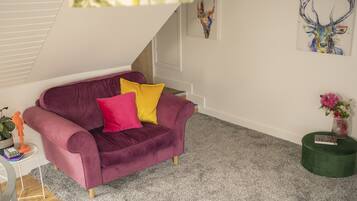 A room with a purple velvet sofa, a small side table with a plant, a green ottoman, and a painting of a deer on the wall.