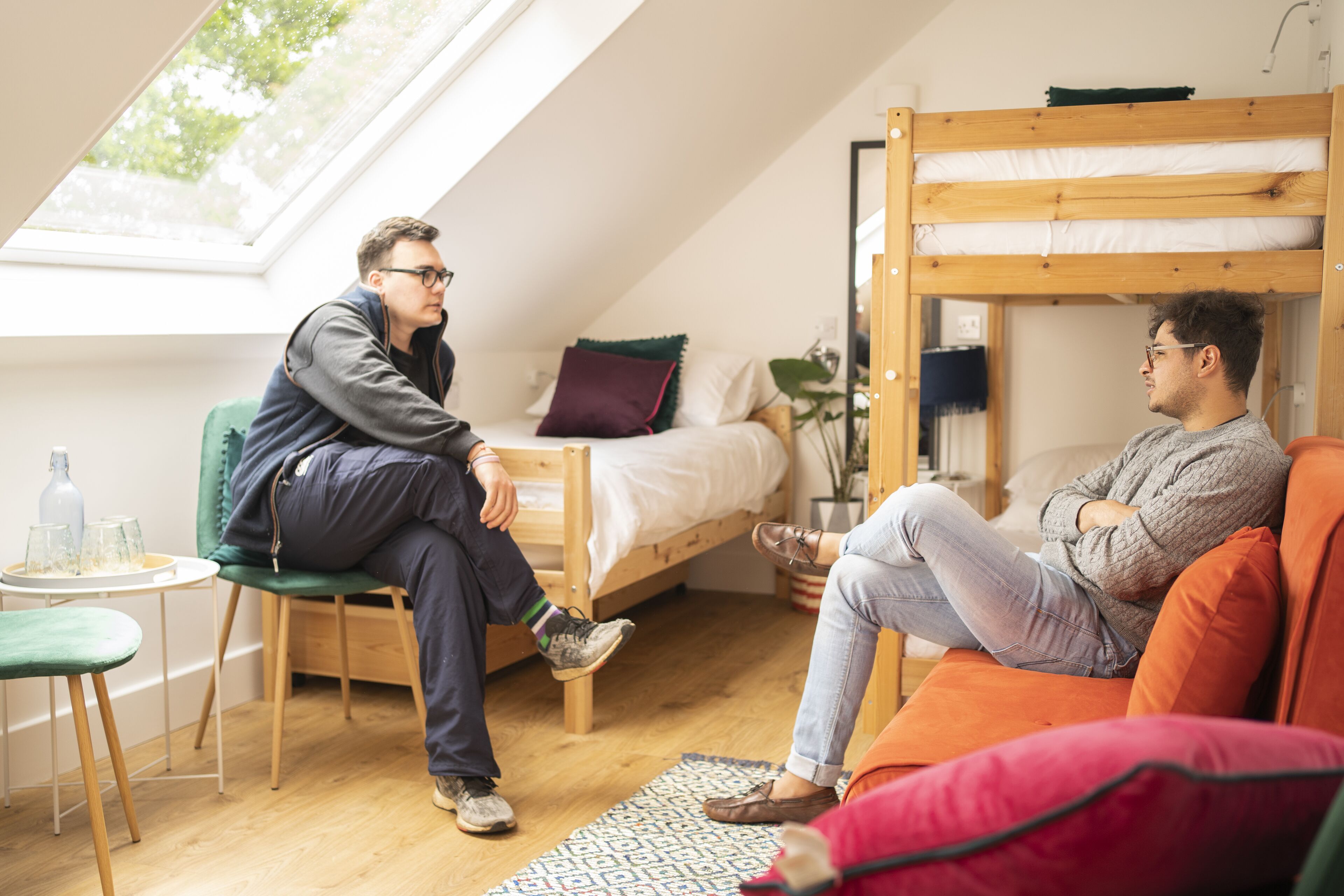 A room with a bunk bed, a green chair, a small table with glasses, and a window with a view of trees.