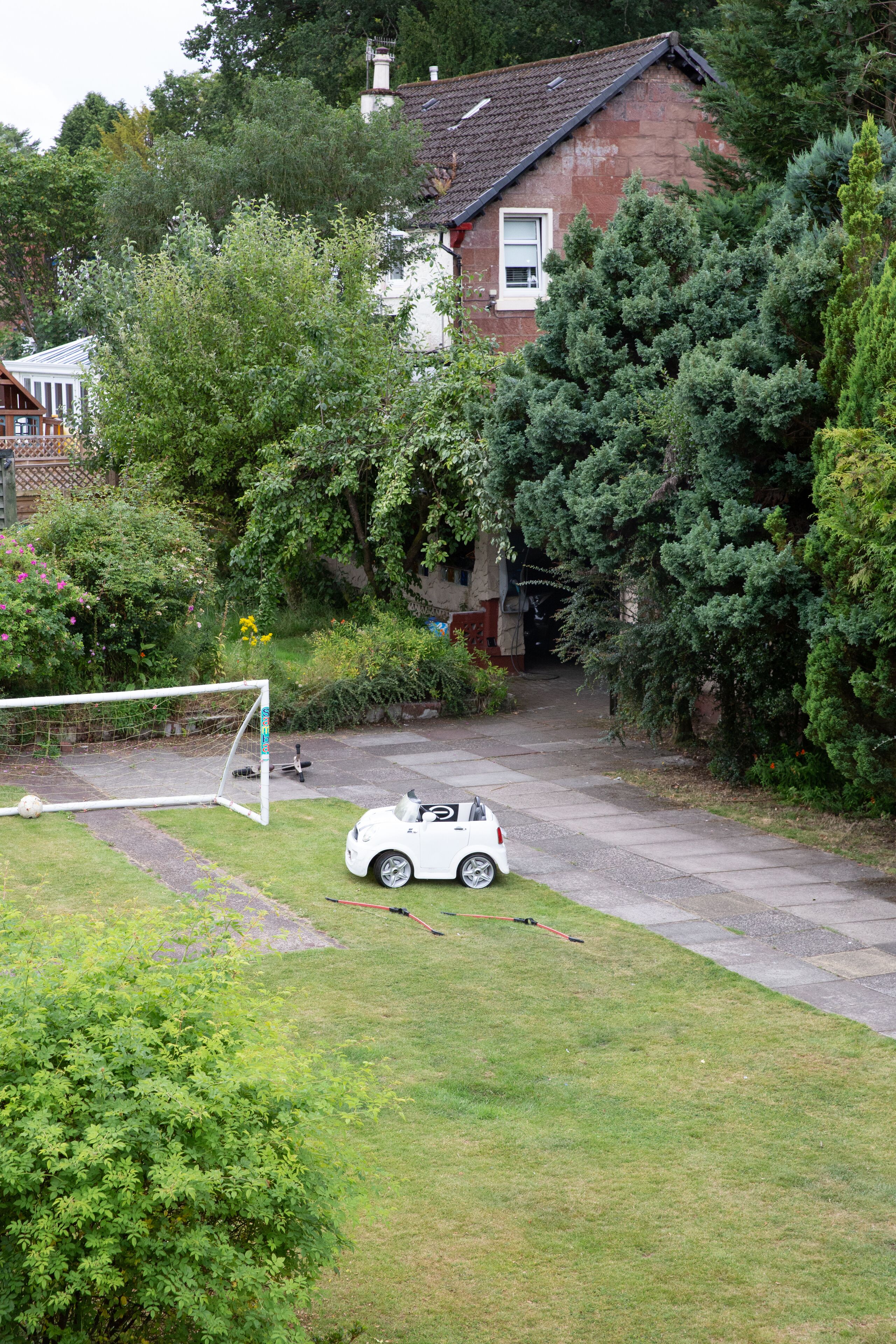 A residential area with a white car parked near a soccer goal, surrounded by lush greenery and a house with a visible chimney.