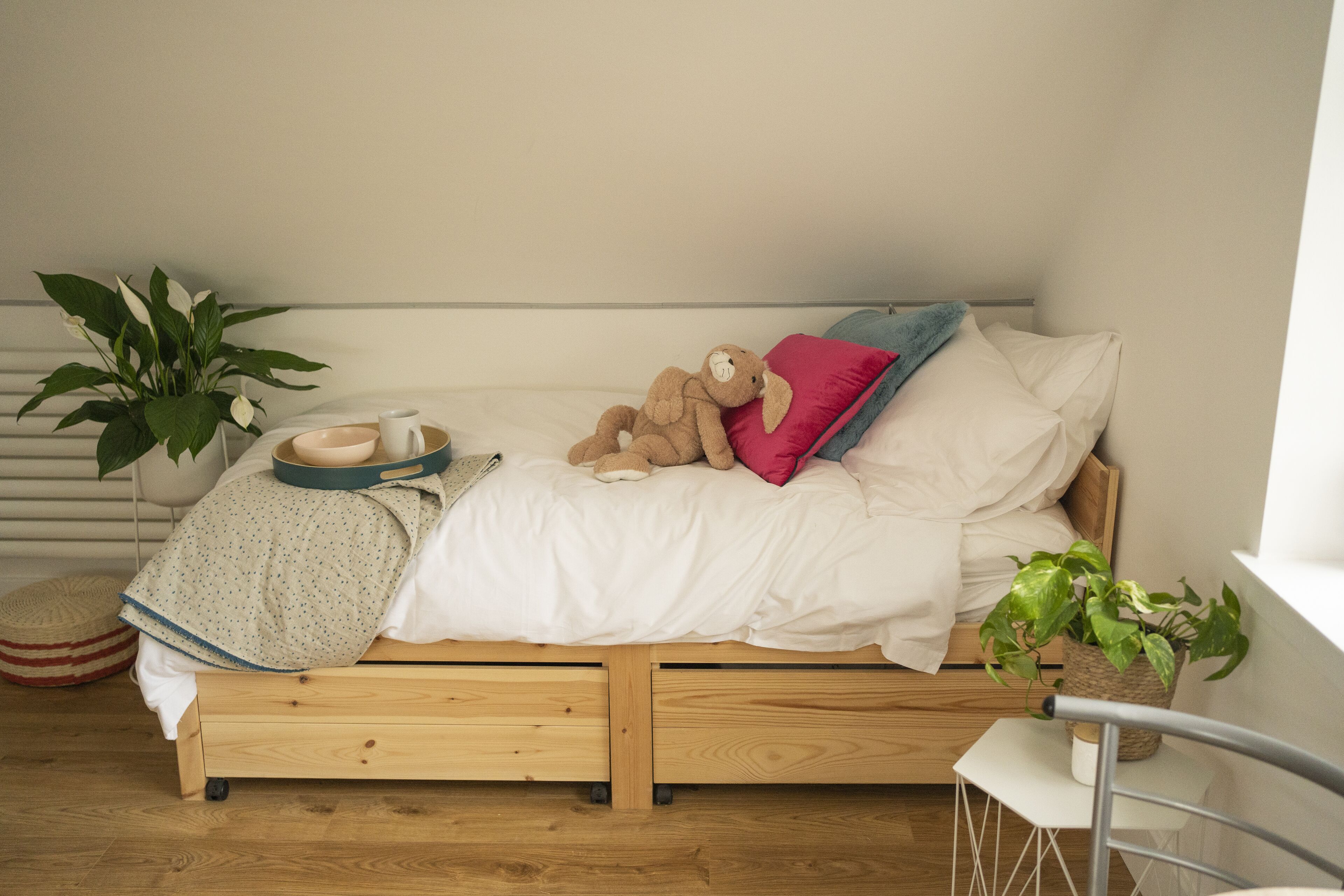 A small wooden bed with a white mattress, a teddy bear, and a potted plant.