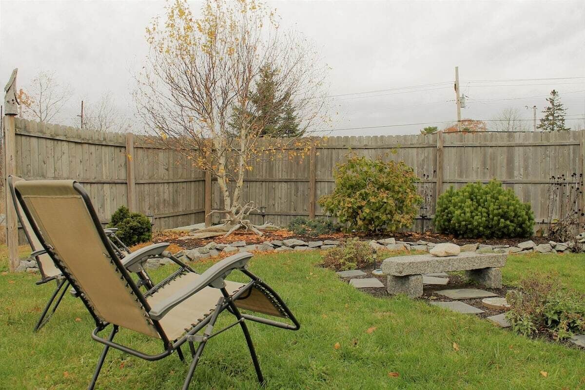A backyard with a wooden fence, a tree, a small bush, and a stone bench.
