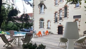 Um deck de madeira com móveis de exterior, uma piscina e um edifício com várias janelas e uma porta.