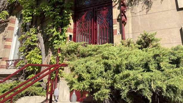 Um edifício com portão de ferro vermelho, varanda coberta com arco decorativo e escadaria que leva à entrada.