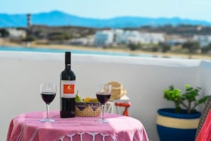 Une table avec une bouteille et deux verres de vin rouge, un panier de fruits et un chapeau sur un balcon donnant sur un paysage côtier.