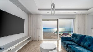 A modern living room with a large flat-screen TV, a blue sofa, a round white coffee table, and a glass door leading to a balcony with a view of the sea. - Villa BAMM with heated 36sqm pool, 5 en-suite bedrooms, and panoramic sea views (Borak)
