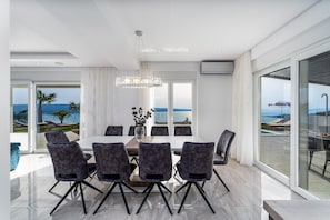 A modern dining area with a large table and chairs, a chandelier, and a view of the sea through large windows.