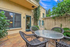 Un patio con mobiliario de mimbre y una mesa de vidrio, rodeado de suelo de piedra y follaje.