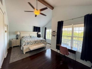 A bedroom with a bed, bedside tables, a desk, a chair, a window with curtains, a ceiling fan, and a view of greenery outside.