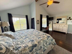 A bedroom with a large bed, a ceiling fan, a dresser, and a window with curtains.