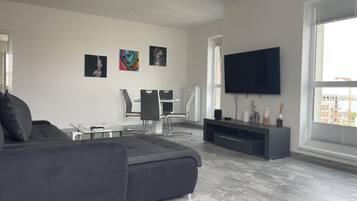 Een moderne woonkamer met een grijze bank, een glazen salontafel, een aan de muur bevestigde televisie en kunstwerken aan de wand.